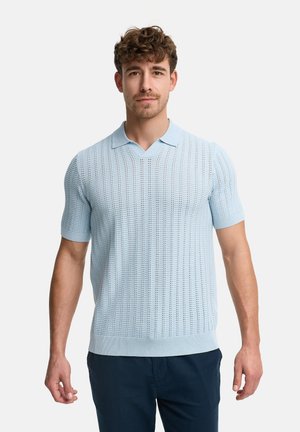 Mand med krøllet hår iført en lyseblå kortærmet strikket polo og mørkeblå bukser, stående mod en ensfarvet hvid baggrund.