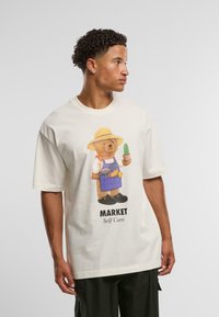 Ung mand med krøllet hår iført en hvid t-shirt med et billede af en bjørn i haveudstyr, der holder en kaktus, med teksten "MARKET Self Care" nedenunder.