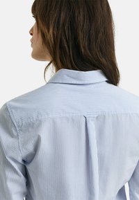 Blusa a righe blu chiaro e bianca con colletto e dettagli di piega sulla schiena, realizzata in un tessuto morbido.