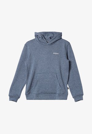 Personne portant un sweat à capuche bleu, un pantalon beige et des tongs noires, debout devant un fond blanc.