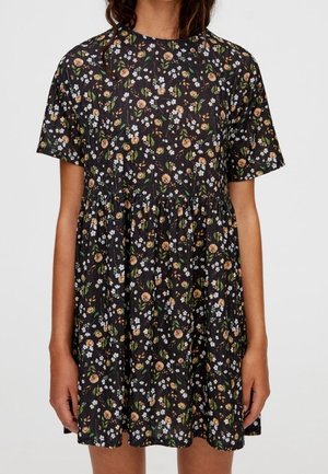 Vrouw draagt een zwart kortärmelig jurkje met een geplooide taille en een bloemenprint van kleine witte en beige bloemen met groene bladeren.