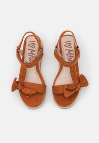 Pair of rust-orange suede sandals with ankle straps and bow accents on front straps, flat sole with light woven trim, viewed from above.