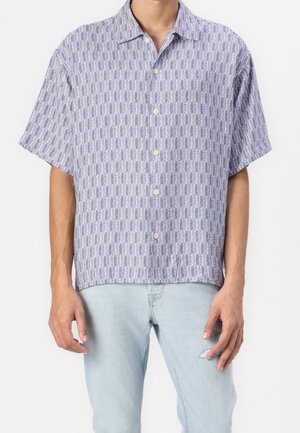 Mand iført kortærmet, knappet skjorte med geometrisk blå og hvidt mønster, kombineret med lyseblå revnede jeans.