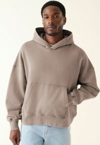 Sudadera marrón de gran tamaño hecha de una tela suave. Presenta un bolsillo frontal, una capucha con cordones y puños y dobladillo acanalados. Se lleva con jeans azul claro.