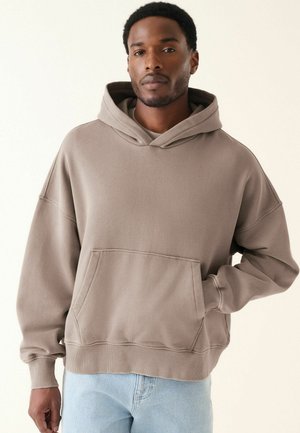Sudadera marrón de gran tamaño hecha de una tela suave. Presenta un bolsillo frontal, una capucha con cordones y puños y dobladillo acanalados. Se lleva con jeans azul claro.