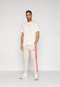 Beżowa koszulka z krótkim rękawem z małym czerwonym logo Adidas, zestawiona z beżowymi spodkami dresowymi z czerwonymi bocznymi paskami i logo Adidas. Białe trampki.