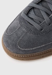 Γκρι σουέντ αθλητικό παπούτσι με στρογγυλερή μύτη, περίπλοκη ραφή και υφασμένο λαστιχένιο πέλμα. Τα επίπεδα γκρι κορδόνια περνούν μέσα από τρύπες.