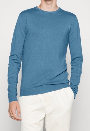 Mand iført en tætsiddende, langærmet blå rundhalset sweater og hvide bukser, med den ene hånd i lommen og en ring på venstre hånd.