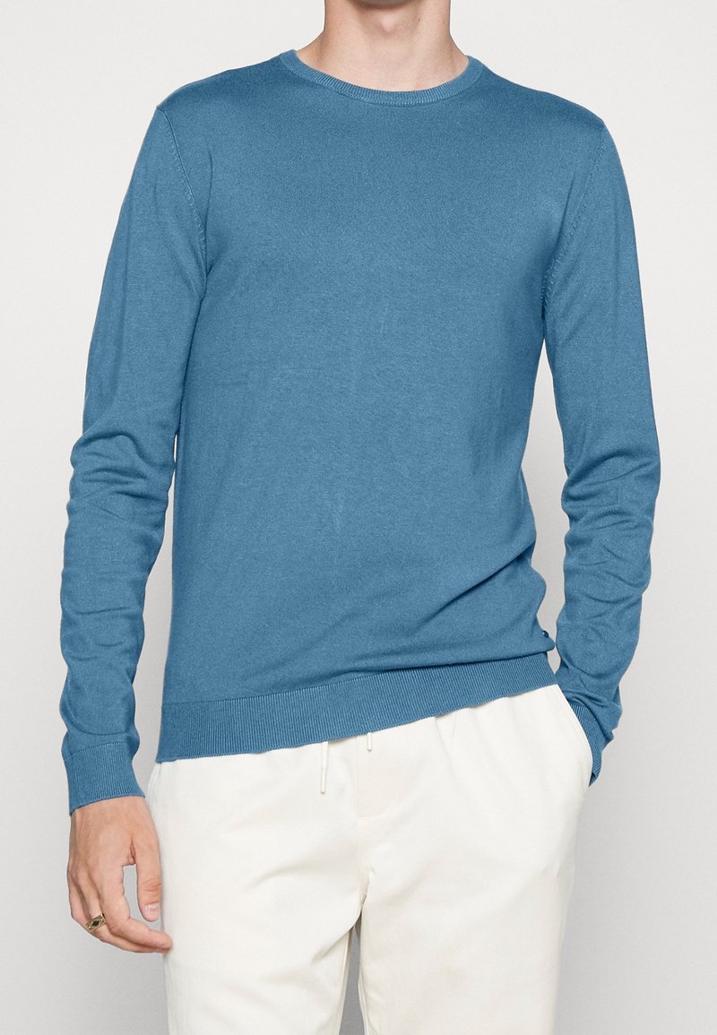 Homme portant un pull bleu à manches longues ajusté avec un col rond et un pantalon blanc, une main dans la poche et une bague à la main gauche.