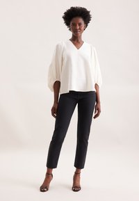 Hvid struktureret bluse med V-hals og pufærmer, parret med sorte tætsiddende jeans og stroppede hæle, mod en neutral baggrund.