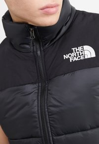 Gilet imbottito nero con colletto alto, chiusura con zip, e logo ricamato bianco "The North Face" sul lato sinistro superiore.