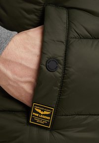 Grüne Pufferjacke mit strukturierter Oberfläche. Verfügt über einen schwarzen runden Knopf und ein rechteckiges Etikett mit gelbem Text und Flügelgrafik.