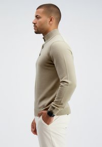 Jersey beige de punto con cuello alto, con tres botones oscuros y puños acanalados. Textura suave, forma ajustada y detalle de logo en el dobladillo.