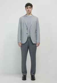 Blazer grigio chiaro con rever a taglio, due bottoni e tasche laterali, abbinato a pantaloni sartoriali coordinati e scarpe nere.