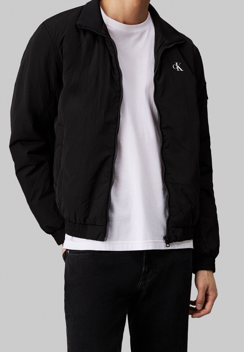 Man som bär en svart zipped jacka med "CK"-logotyp på bröstet, över en vit t-shirt och svarta jeans, stående mot en grå bakgrund.