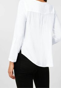 Camicia bianca a maniche lunghe con orlo arcuato e dettagli di pieghe sul retro, abbinata a jeans neri aderenti, che evidenziano una texture di tessuto liscia.