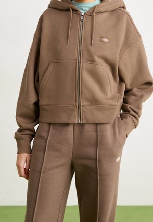 Persona que lleva una sudadera con capucha marrón con cremallera y pantalones de chándal marrones a juego con pequeños logos de la marca, de pie con una mano en el bolsillo.