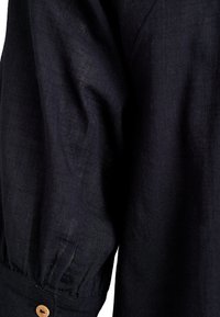 Detail tmavomodrej ľanovej košele, zobrazujúci jemnú textúru, mierny lesk, nariasený rukáv a drevený gombík na manžete.