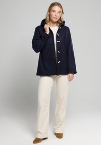 Cappotto navy con cappuccio e bottoni in legno, dotato di tasca frontale e vestibilità rilassata. Abbinato a pantaloni crema e stivali alla caviglia marroni.