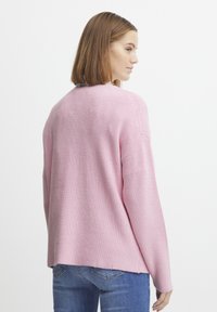 Svetlo roza rebrast pulover s sproščenim krojem, ki ima okroglo izrez in spuščena ramena. Nosite ga z modrimi kavbojkami, prikazan od zadaj.