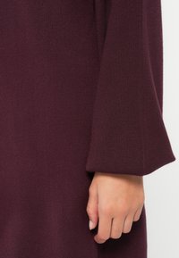 Abito intrecciato bordeaux con maniche lunghe, caratterizzato da una vestibilità comoda e una texture a coste. Dettaglio ravvicinato della manica e della mano.