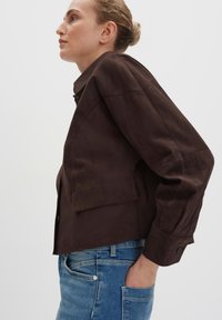 Braune Wildleder-Cropped-Jacke mit hohem Kragen, sichtbaren Nähten und einem Ein-Knopf-Verschluss, kombiniert mit hellblauen Jeans.