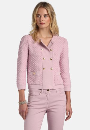 Žena oblečená v textúrovanom svetloružovom dvojradovom saku so zlatými gombíkmi a zodpovedajúcich nohaviciach, doplnená perlovým náramkom.