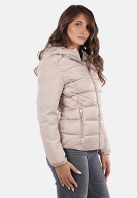 Giacca imbottita color beige chiaro con cappuccio, motivo a rombi, tasche con zip e hardware color oro, caratterizzata da una texture morbida e liscia.