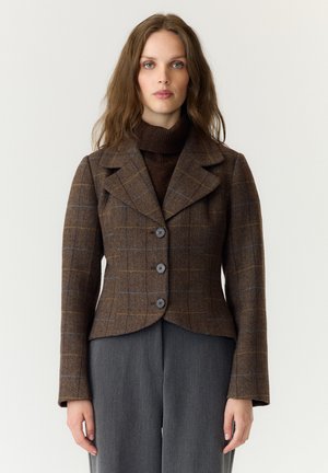 Kvinde iført en brun ternet blazer over en mørkebrun rullekrave og grå bukser, stående mod en ensfarvet lys baggrund.