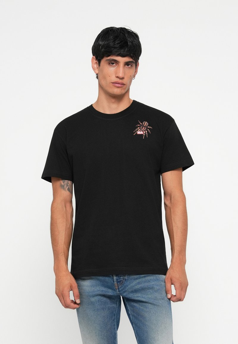 T-shirt en coton noir avec un graphisme d'araignée rouge et orange sur la poitrine gauche. Encolure ras du cou classique et manches courtes ; design minimal.