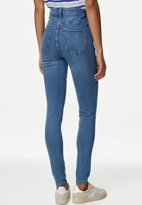 Vaqueros de mezclilla azules ajustados con cinco bolsillos, cintura alta y ligero estiramiento. Llevados con zapatillas de deporte de bajo corte y color claro.