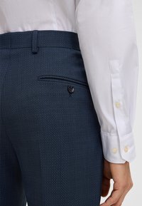 Dunkelblaue gemusterte Hosen aus glattem Stoff, mit einer Gesäßtasche mit Knopf und einem Gürtelschlaufen am Bund. Weiße Hemdmanschetten sind sichtbar.