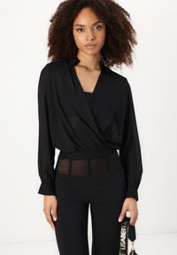Blusa nera avvolgente in tessuto trasparente con maniche lunghe, abbinata a pantaloni neri a vita alta con pannello in rete.