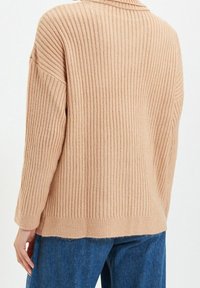 Beżowy sweter dziany w prążki o luźnym kroju, z kołnierzykiem i długimi rękawami. Dolny brzeg jest lekko przetarty, co dodaje tekstury.