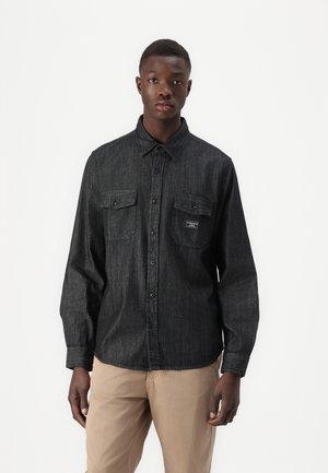Dark denim shirt with long sleeves, button-down collar, two chest pockets, and a small logo patch. Smooth texture, modern fit.