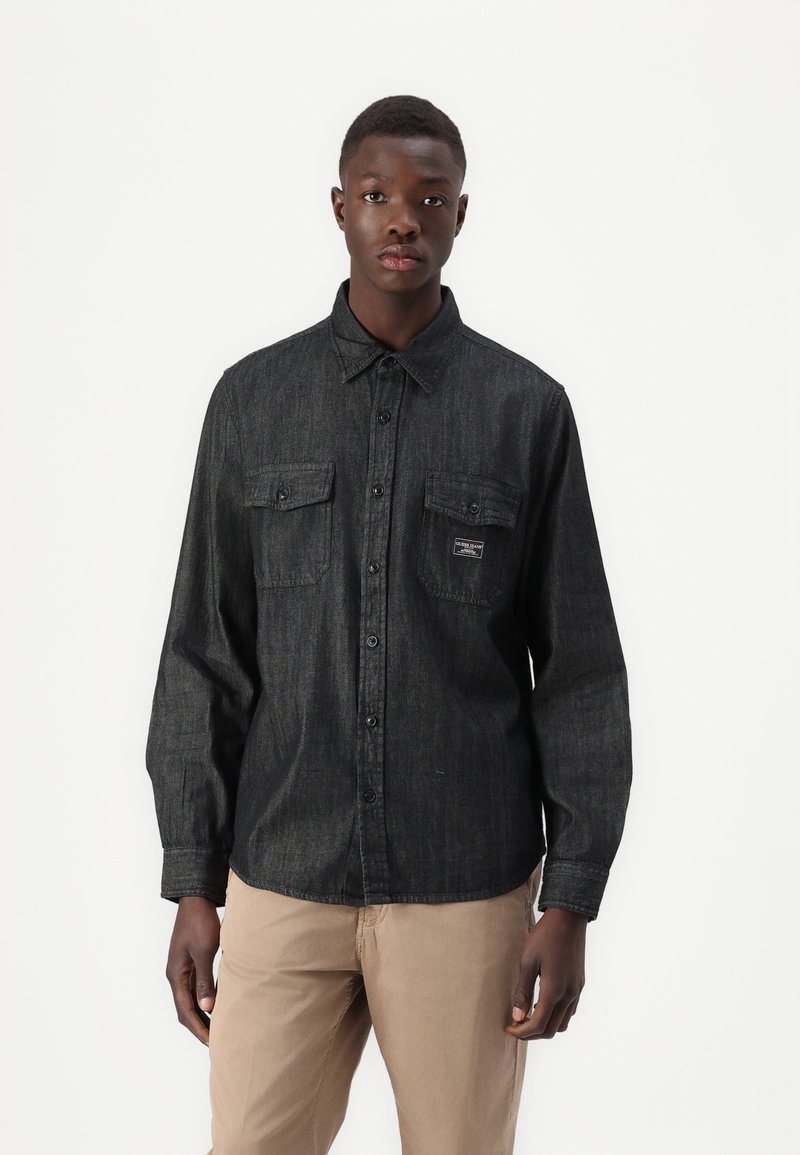 Dark denim shirt with long sleeves, button-down collar, two chest pockets, and a small logo patch. Smooth texture, modern fit.