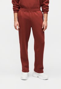 Personne portant un pantalon de survêtement rouge et un sweat-shirt assorti avec des baskets blanches, debout contre un fond clair uni.