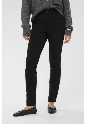 Personne portant un pantalon slim noir, un pull en maille gris à manches longues et des ballerines noires, debout sur un fond blanc.