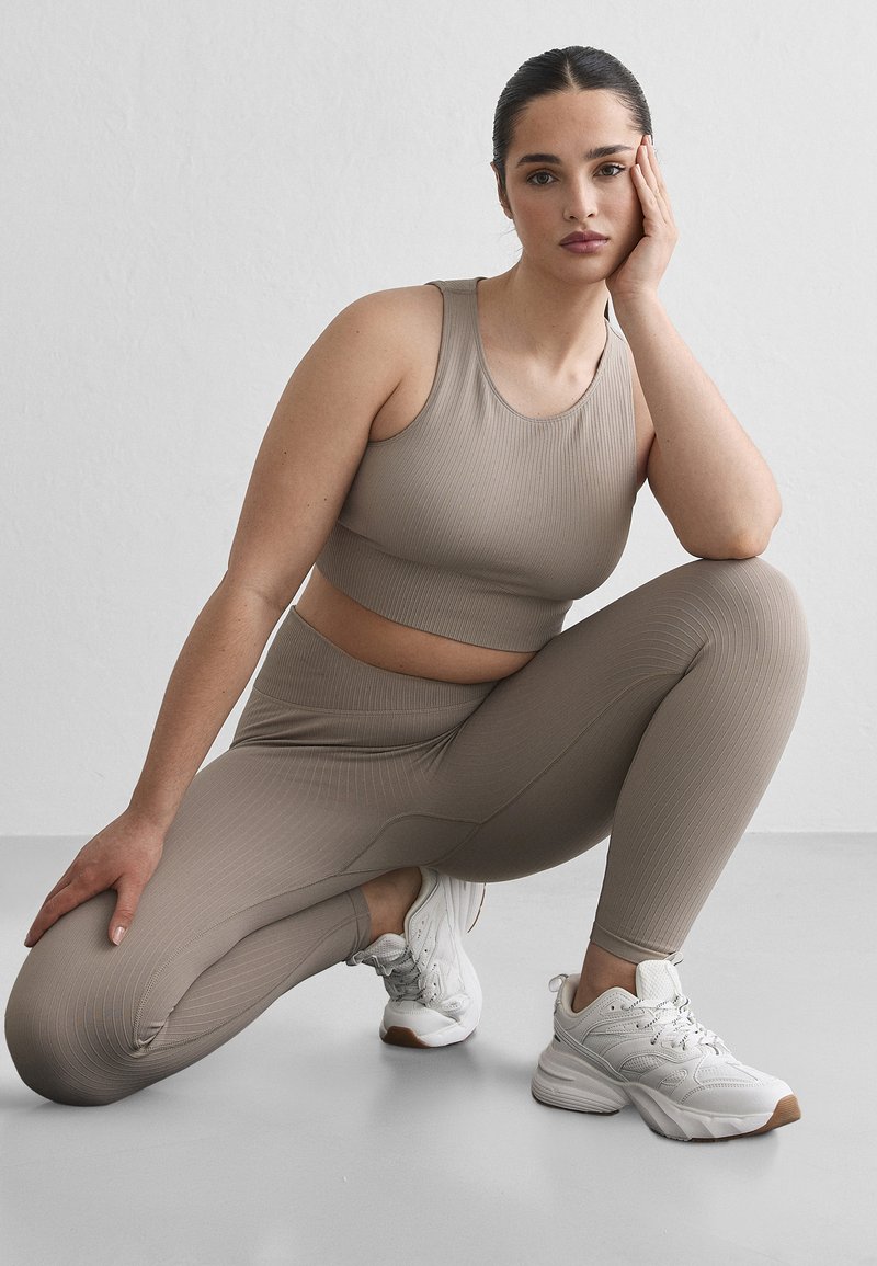 Femme en tenue de sport beige côtelée et baskets blanches, agenouillée sur une jambe, reposant son visage sur sa main sur un fond gris clair uni.