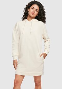 Gaiši bēšas kapuces sporta kleitas ar garām piedurknēm, sānu kabatām, ribotu aproču apdari un brīvu pieguļošos ar taisnu apakšmalu.