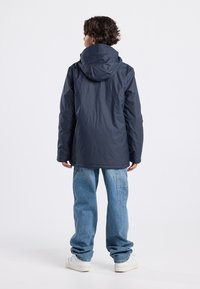 Navy blue waterproof jacket with a hood, featuring a smooth texture and a relaxed fit. Paired with light blue baggy jeans and white sneakers.