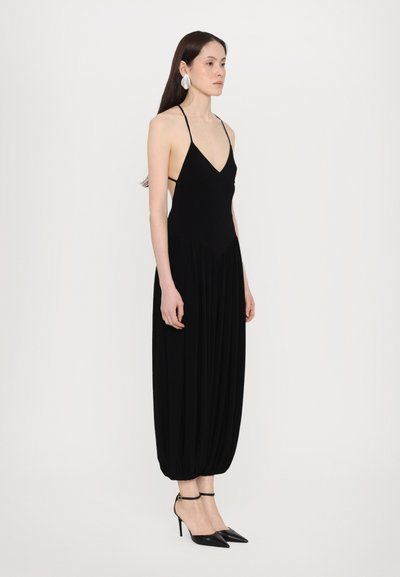 Schwarzer Jumpsuit mit dünnen Trägern, tiefem V-Ausschnitt und gerafftem Beinsaum. Komplementiert von spitzen schwarzen Heel und großen silbernen Ohrringen.