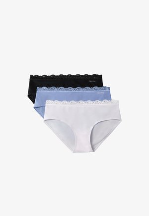 Femme debout pieds nus devant un fond gris, portant un soutien-gorge rose clair et une culotte assortie, les bras croisés et une main touchant ses cheveux.