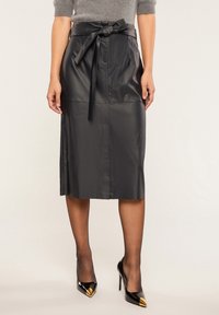 Musta nahkainen midi-hame, jossa on sidottu vyötärö, sileä pinta ja sivuaukot liikkuvuuden helpottamiseksi, yhdistettynä terävin kärkin korkeisiin kenkiin.