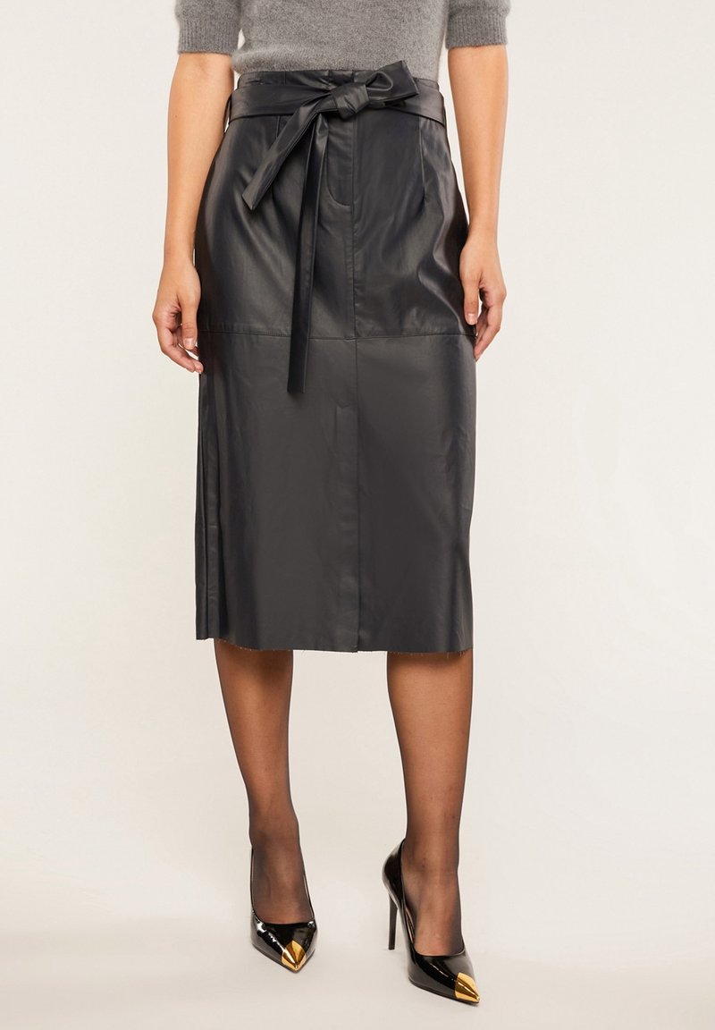 Musta nahkainen midi-hame, jossa on sidottu vyötärö, sileä pinta ja sivuaukot liikkuvuuden helpottamiseksi, yhdistettynä terävin kärkin korkeisiin kenkiin.