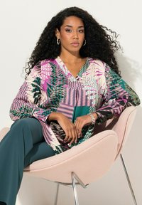 Blusa a maniche lunghe colorata con un vivace mix di viola, verde e texture a fantasia. Abbinata a pantaloni turchesi, la modella è seduta su una sedia rosa.