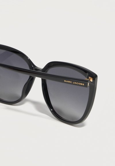 Μαύρα υπερμεγέθη γυαλιά ηλίου με τετράγωνο σχήμα, φακούς με βαθμιαία σκουρότητα και χρυσή επιγραφή "MARC JACOBS" στο πλαϊνό μπράτσο.