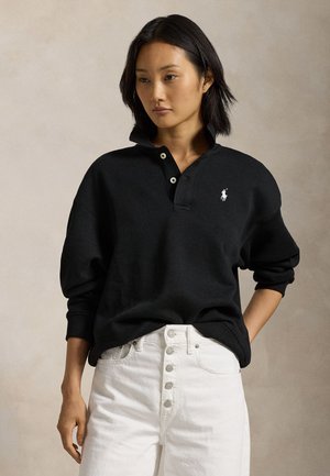 Γυναίκα με μαύρο φούτερ με γιακά και λευκό τζιν με κουμπιά, στέκεται απέναντι σε απλό μποέζ φόντο, με το ένα χέρι στην τσέπη.