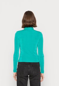 Top ajustado de manga longa em turquesa brilhante, com textura suave. Apresenta um colarinho alto e mangas finas, combinado com calças pretas.