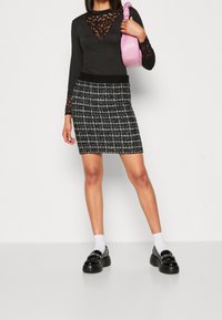 Černý top s dlouhými rukávy a krajkovými detaily, doplněný černobílou kostkovanou minisukní. Modelka drží růžovou kabelku a má na sobě černé mokasíny.